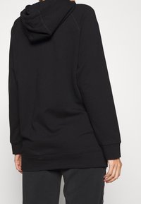 Sudadera negra con un corte holgado, mangas largas y capucha con cordón. Tela suave con puños y dobladillo acanalados. Costuras mínimas.