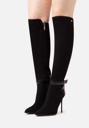 Zwarte suède kniehoge laarzen met spitse neuzen, naaldhakken, zijrits en enkelriem met metalen gesp en hangend kettingbedeltje.