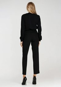 Czarna bluzka z długim rękawem w połączeniu z dopasowanymi czarnymi spodniami, o slim fit, gładkiej fakturze i czystej sylwetce. Całość uzupełniają wysokie obcasy.