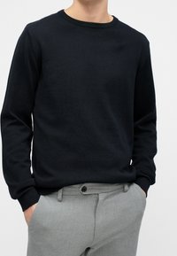 Black knit sweater with a crew neckline, ribbed cuffs, and relaxed fit, paired with light gray trousers featuring a button and belt loops.