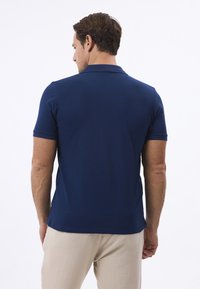 Tmavě modrá polo košile s volným střihem, krátkými rukávy a límcem. Vyrobena z hladkého materiálu, má rovný lem a minimální designové detaily.
