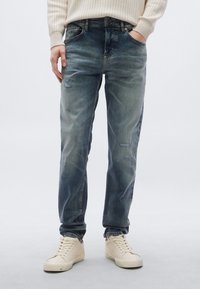 Világoskék, kopott hatású farmernadrág, enyhén szakadt részletekkel és egyenes szabással. Krémszínű tornacipővel párosítva.