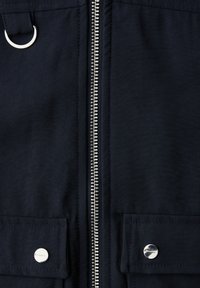 Metallinen vetoketju ja hopea rengas tummassa kankaassa, jossa on kaksi läppätaskua, jotka on suljettu hopeisilla neppareilla.