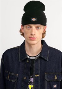 Sort strikket hue med et logo, båret med en mørk denimjakke med gul syning og knapper, over en sort mesh og grafisk skjorte.