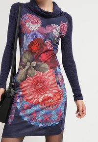 Blommönstrad klänning med cowl-krage, som har mörkblå långa ärmar. Framsidan är prydd med livliga blommor i olika färger och en figurnära design.