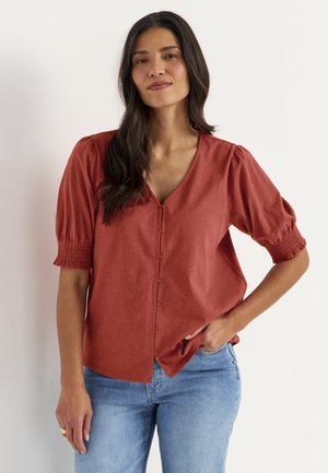 Vrouw staat met hand in zak, draagt een roestkleurige blouse met knopen en pofmouwen en lichtblauwe jeans tegen een witte achtergrond.