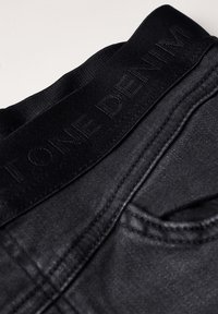 Svarta denimjeans har en texturerad midja med präglad logotyp, kontrasterande sömmar och en liten bakfickdetalj.