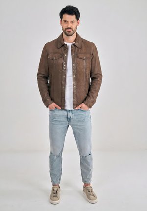 Muž v hnědé kožené bundě, bílém tričku, světlých roztrhaných džínách a béžových teniskách stojí s rukama v kapsách.