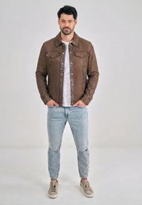 Mand iført brun læderjakke, hvid t-shirt, lyse revnede jeans og beige sneakers, stående med hænderne i lommerne.