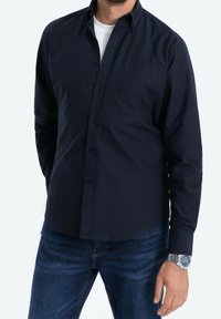 Tmavě modrá košile na knoflíky s dlouhými rukávy, jednou náprsní kapsou a hladkou texturou, doplněná o džíny a stříbrné hodinky.