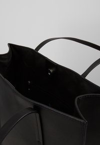 Zwarte leren tote bag met een gladde textuur, open bovenkant en dubbele handvatten. Interieur beschikt over een ritszak en een magnetische sluiting.