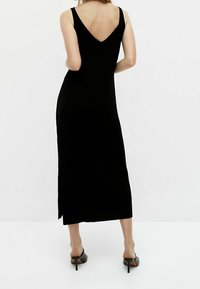 Schwarzes ärmelloses Maxikleid mit V-Ausschnitt, das eine figurbetonte Silhouette, seitlichen Schlitz und weichen Stoff aufweist; kombiniert mit schwarzen hohen Schuhen.