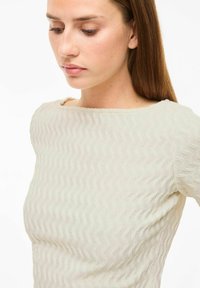 Top texturizado color ecru con un patrón ondulado y un escote barco. La tela parece suave y ligeramente transparente. El diseño ajustado realza la silueta.
