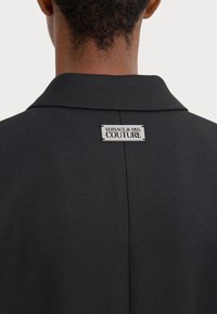 Crni blejzer sa strukturiranim kragnom, sa srebrnom oznakom "Versace Jeans Couture" blizu vrata. Tekstuirana tkanina sa krojem koji dobro pristaje.