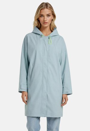 Femme aux cheveux blonds portant un imperméable à capuche bleu clair à manches longues et un jean bleu clair, debout devant un fond uni.