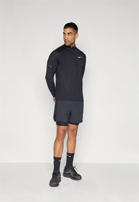 Svart långärmad sporttröja med dragkedja, matchad med mörka shorts, svarta strumpor och sneakers. Logotyp synlig på bröstet och skorna.