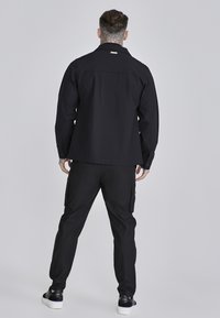 Chaqueta negra con un corte relajado y puños abotonados, combinada con pantalones cargo a juego que cuentan con bolsillos laterales, todo ello sobre un fondo neutro.