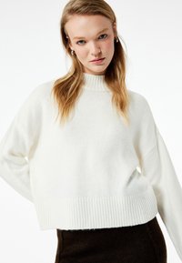 Suéter blanco de punto con cuello alto y dobladillo acanalado, con un ajuste relajado y hombros caídos, colocado sobre un fondo blanco liso.