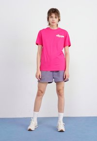 T-shirt rose à manches courtes avec logo, associé à un short gris. Le mannequin se tient sur un sol bleu devant un fond blanc, portant des baskets blanches.