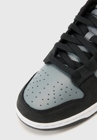 Fekete velúrból és szürke bőrből készült sneaker. Jellemzője a perforált orr-rész, fekete fűzők és egy fehér középtalp, amelyhez egy texturált fekete talp tartozik.