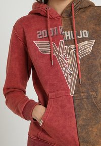 Czerwona i brązowa bluza z kapturem w blokowe kolory z nadrukiem graficznym. Wykonana z miękkiej bawełny, posiada przednią kieszeń, sznurki do ściągania i ściągacze na rękawach.