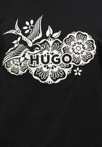 Λευκό κεντητό σχέδιο με λουλούδια και πουλί που περιβάλλει τη λέξη «HUGO» σε μαύρο ύφασμα.
