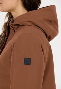 Bruine waterdichte jas met capuchon, gestructureerd materiaal, verstelbare trekkoord en een zwart logo op de mouw.