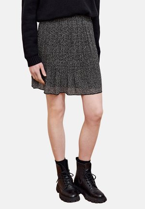 Person trägt ein schwarzes Langarmshirt, einen plissierten Minirock mit Leopardenmuster und schwarze Schnür-Stiefel vor einfachem Hintergrund.