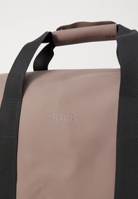 Hnedý vodoodolný taška cez rameno s čiernymi nylonovými popruhmi a embosovaným logom "RAINS". Má hladký, matný povrch a pevné kovové akcenty.
