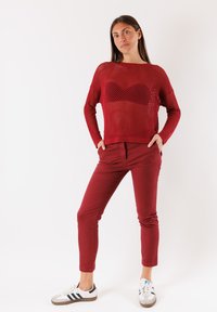 Maglione a rete rosso con scollatura ampia, abbinato a pantaloni rossi aderenti e sneakers bianche con strisce nere. Outfit semplice e casual.