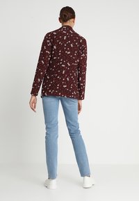 Bordowa marynarka w kwiatowy wzór z długimi rękawami, zestawiona z jasnoniebieskimi obcisłymi dżinsami i białymi trampkami, widziana od tyłu.