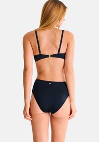 Femme aux longs cheveux blonds portant un maillot de bain noir deux pièces, vue de dos sur un fond clair uni.