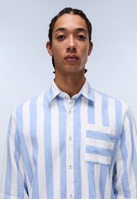 Camisa de lino a rayas en azul claro y blanco, con un cierre de botones, un bolsillo en el pecho con rayas horizontales y un diseño de cuello clásico.