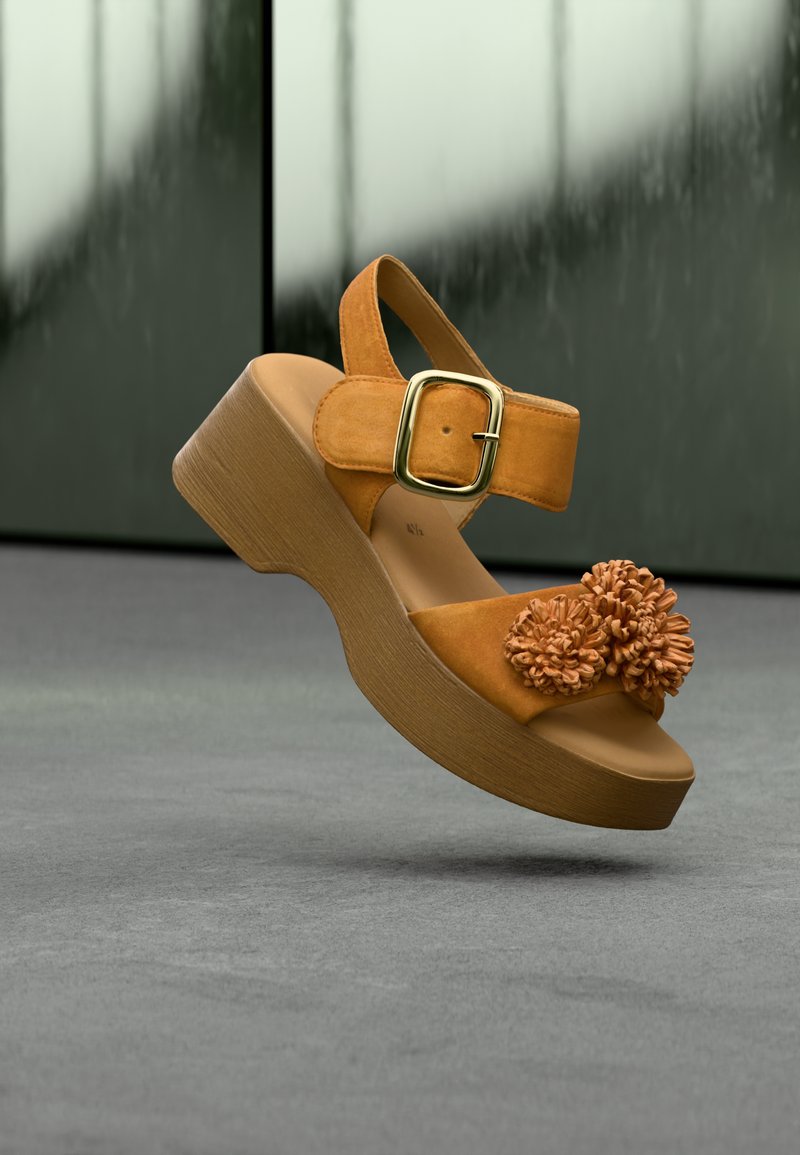 Πλατφόρμα σανδάλι από μουσταρδί σουέντ, με φαρδιά λουράκι με χρυσή αγκράφα και διακοσμητικά με λουλούδια μπροστά, τοποθετημένο σε ξύλινη σόλα.