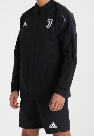 Osoba ubrana w czarną, długorękawową kurtkę Adidas Juventus oraz czarne spodenki Adidas, stojąca z rękami swobodnie po bokach.