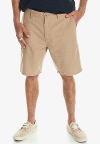 Beige shorts i ett texturerat vävt tyg, med knappstängning fram och två sidofickor. Bärs tillsammans med ljusfärgade sneakers.