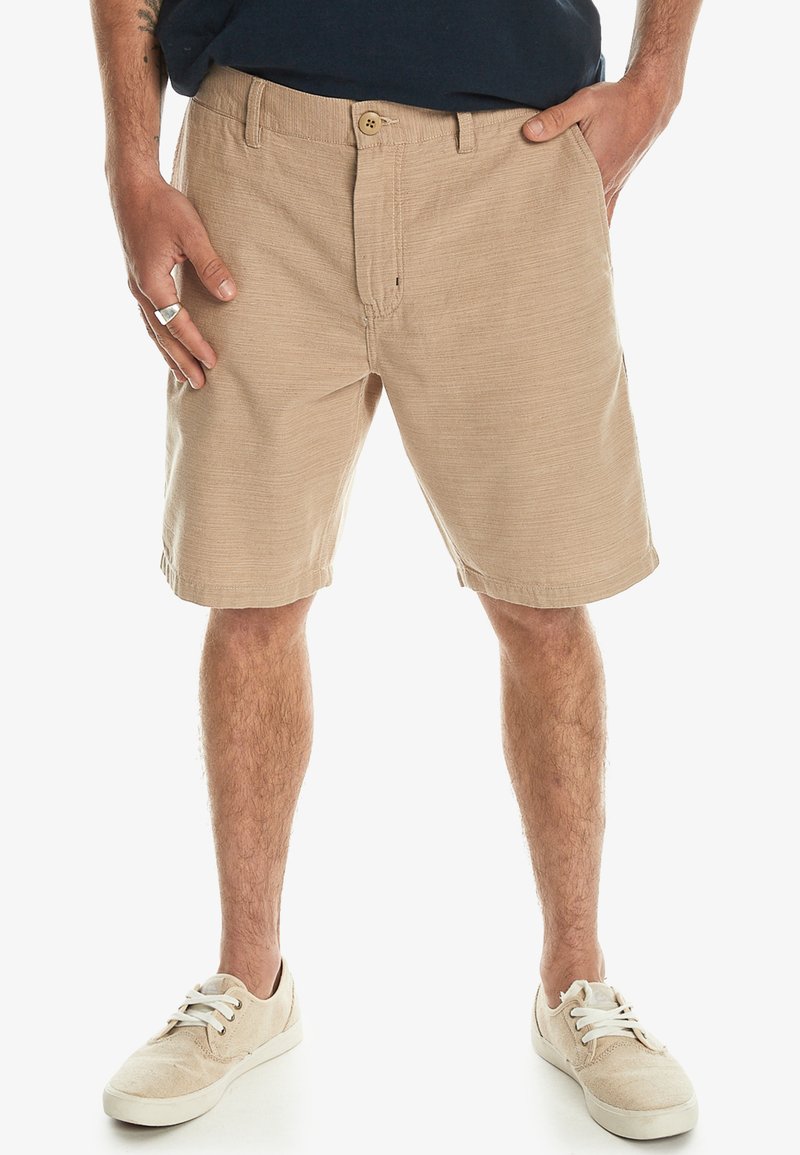 Beige shorts i ett texturerat vävt tyg, med knappstängning fram och två sidofickor. Bärs tillsammans med ljusfärgade sneakers.