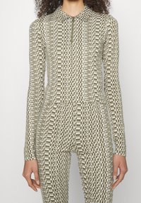 Långärmad tröja med dragkedja och matchande tights i olivgrönt med krämfärgat rutmönster och upprepat textmotiv över hela plagget.