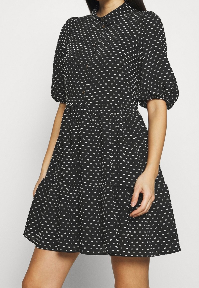 Femme portant une robe courte noire avec de petits motifs carrés blancs, des manches courtes bouffantes et des boutons à l'avant.
