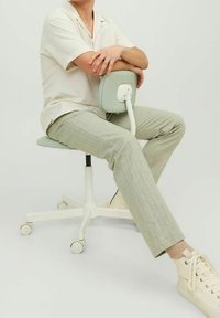 Arbetsstol med en ljusgrön vadderad sits och ryggstöd, vit metalbas och hjul. Person sitter, klädd i en ljus skjorta och beiga byxor.