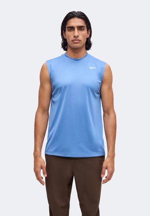Homme aux cheveux mi-longs et foncés portant un débardeur sportif bleu clair et un pantalon marron, debout devant un fond clair uni.