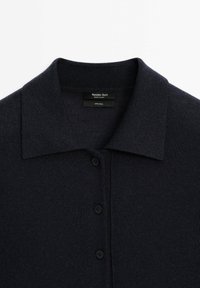 Tummansininen villa-polosukot, jossa on korkea kaulus, viisi mustaa nappia ja pehmeä rakenne. Tunnetta näyttävä etiketti osoittaa 100 % villakokoonpanon.