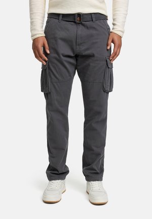Homme portant un pantalon cargo gris foncé avec des poches latérales, des baskets blanches et une chemise légère à manches longues, debout devant un fond uni.