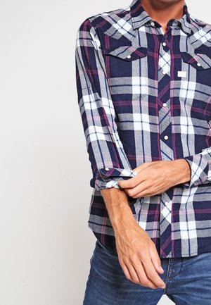 Homme retroussant les manches d'une chemise à carreaux bleu marine, blanc et rouge, portant un jean bleu, sur un fond clair uni.
