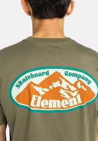 Olivovo zelené tričko s krátkymi rukávmi s veľkým logom horského motívu, s nápisom „Element“ v bielej farbe a „Skateboard Company“ s akcentmi v zelenej a oranžovej farbe.