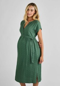 Groene zwangerschapsjurk met korte mouwen, V-hals, strikdetail bij de taille en een knie-lange zoom. Heeft een stippenpatroon door het geheel.