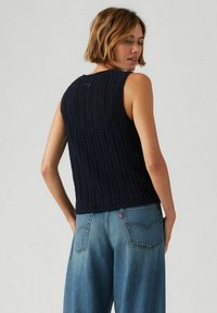 Mörkblå stickad ärmlös topp med texturerat mönster; har en rundad halsringning och ribbad nederdel. Bärs med ljusblå vida jeans.