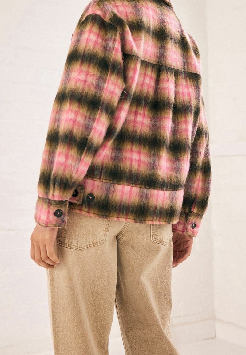 Personne portant une veste à carreaux rose, marron et jaune avec des poignets boutonnés et un pantalon beige, photographiée de dos.