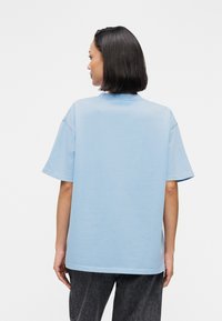 Γυναίκα με κοντά σκούρα μαλλιά, που φοράει ένα άνετο φωτεινό μπλε T-shirt και μαύρο παντελόνι, εμφανίζεται από πίσω μπροστά σε ένα απλό λευκό φόντο.