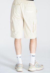 Beige cargo shorts med elastisk talje og flere lommer, med trykt branding på bagsiden. Bæres med hvide sokker og sneakers.